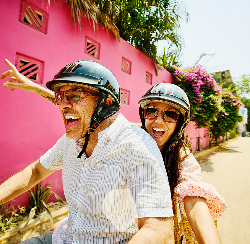Couple in their 40's riding a scooter through a colorful neighborhood on a sunny day