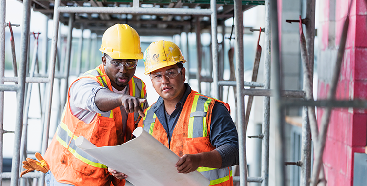 Two construction workers in hardhats and safety vests discuss construction plans