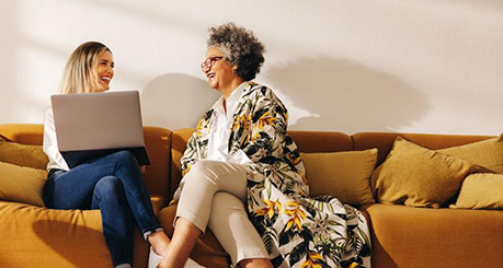 Two smiling friends sit together on a couch, one holds open laptop