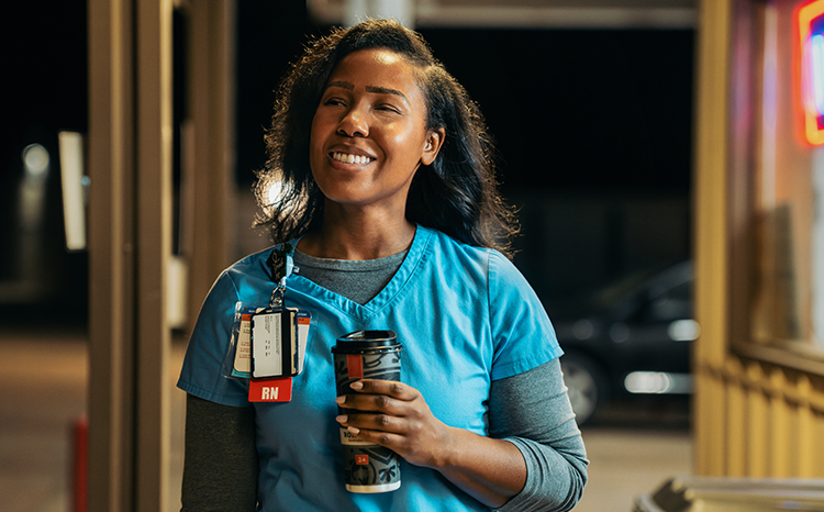 Nurse in blue scrubs holding coffee looks away and smiles