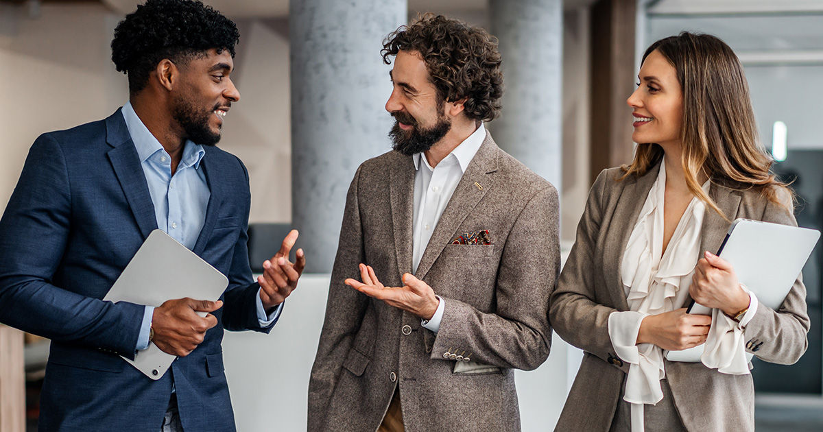 Three professionals walking and talking in an office, holding tablets and documents.