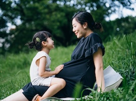 Adult sitting on a blanket in a grassy field with a child seated nearby, outdoors in a park-like setting.